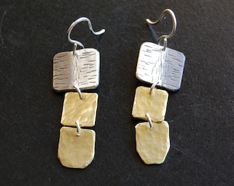 Op de afbeelding: Een paar zilver- en goudkleurige oorbellen. Elke oorbel heeft drie vierkante vormen die met kleine lussen zijn verbonden. De bovenste vierkant is zilverkleurig met een textuurontwerp, terwijl de onderste twee goudkleurig zijn met een gehamerde afwerking.