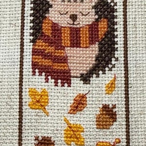 May include: Cross-stitch bookmark depicting a hedgehog with a striped scarf, surrounded by autumn leaves in yellow, orange, and brown. The bookmark has a brown border and is set against a neutral background.