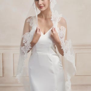 Op de afbeelding: Een witte bruidssluier met delicate kanten rand. De sluier is gedrapeerd over een witte satijnen jurk met een V-hals en een hoge split. De kanten details voegen een vleugje elegantie toe aan het bruidsensemble.
