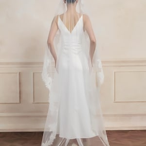 Op de afbeelding: Een witte trouwjurk met een V-hals en een lange, vloeiende sluier. De sluier heeft een delicate kanten rand langs de randen. De bruid staat met haar rug naar de camera en toont de jurk en sluier.