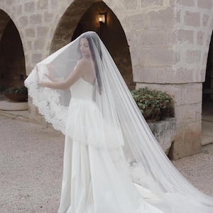 Op de afbeelding: Een witte strapless trouwjurk met een lange, vloeiende sleep. De bruid draagt een doorschijnende sluier met kanten rand. De achtergrond toont stenen bogen en een binnenplaats.