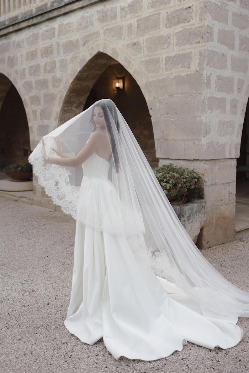 Op de afbeelding: Een witte strapless trouwjurk met een lange, vloeiende sleep. De bruid draagt een doorschijnende sluier met kanten rand. De achtergrond toont stenen bogen en een binnenplaats.