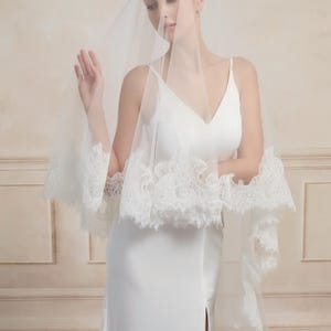 Op de afbeelding: Een bruid draagt een witte trouwjurk met een V-hals en een lange, zwierige rok. Ze draagt ook een doorschijnende, witte sluier met delicate kanten randen. De sluier bedekt haar schouders en armen.