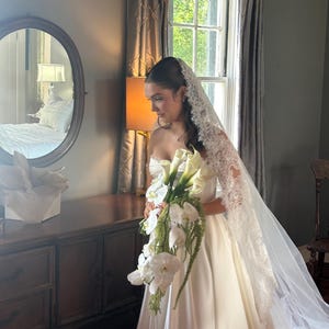 Op de afbeelding: Een bruid in een witte strapless trouwjurk houdt een boeket witte bloemen vast. Ze draagt een lange sluier met kanten rand. De sc&egrave;ne speelt zich af in een kamer met een donkere houten dressoir en een raam met grijze gordijnen.