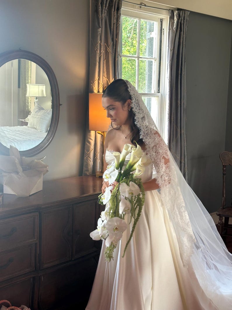Op de afbeelding: Een bruid in een witte strapless trouwjurk houdt een boeket witte bloemen vast. Ze draagt een lange sluier met kanten rand. De sc&egrave;ne speelt zich af in een kamer met een donkere houten dressoir en een raam met grijze gordijnen.
