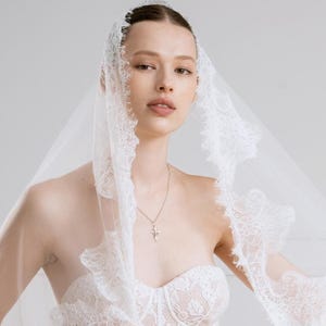 Op de afbeelding: Een witte kanten trouwjurk met een sweetheart halslijn en een bijpassende sluier. De sluier is afgezet met delicate kant en omlijst het gezicht. Een zilveren kruis hanger hangt aan een fijne ketting.