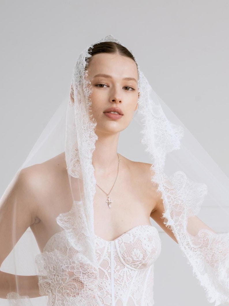 Op de afbeelding: Een witte kanten trouwjurk met een sweetheart halslijn en een bijpassende sluier. De sluier is afgezet met delicate kant en omlijst het gezicht. Een zilveren kruis hanger hangt aan een fijne ketting.