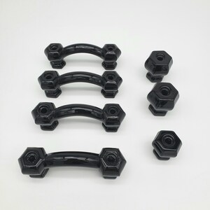 Puede incluir: Un conjunto de herrajes negros para armarios, que incluye cuatro tiradores curvos y tres pomos hexagonales. Los tiradores tienen un diseño curvo con un agarre central y extremos hexagonales. Los pomos también son hexagonales y tienen un acabado brillante.