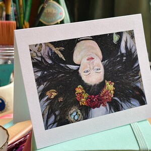 May include: A greeting card featuring an illustration of a woman with long black hair, a floral crown, and blue eyes. The artwork includes a nest with blue eggs and autumn leaves. The card is displayed on a mint green surface.
