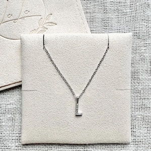 Könnte beinhalten: Eine silberne Kettenkette mit einem kleinen, diamantbesetzten Buchstaben L Anhänger. Die Kette wird auf einem weißen Samt Hintergrund präsentiert.
