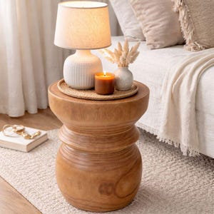 May include: A wooden side table with a white lamp, a candle, and a small vase with dried flowers. The lamp has a beige lampshade. The table is round and has a natural wood finish. A woven tray holds the candle and vase.