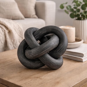 May include: A dark gray decorative knot sculpture made of wood. The intertwined loops create a unique, modern design. The sculpture is displayed on a wooden coffee table, suggesting a home decor theme.