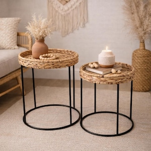 May include: Two round side tables with woven rattan tops and black metal legs. One table holds a terracotta vase with dried flowers and a wooden bead garland. The other table has a candle and a stack of books.