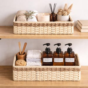 May include: Two woven wicker baskets on a wooden shelf. The top basket holds rolled towels, a candle, and decorative items. The bottom basket contains shampoo, conditioner, and body wash bottles, along with a loofah and wooden utensils. The baskets have a natural color.