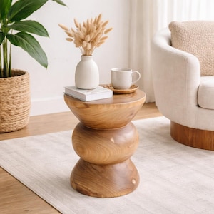 May include: A wooden side table with a unique hourglass shape. The table is made of light brown wood and has a smooth finish. A white vase with dried flowers, a book, a mug, and a coaster are on top of the table. A potted plant and a cream-colored armchair are in the background.