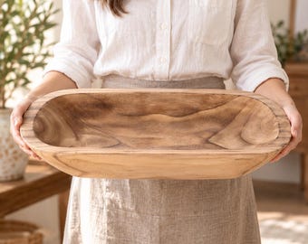 May include: An elongated, oval-shaped wooden dough bowl featuring a natural light brown finish with visible dark wood grain patterns. The bowl has a smooth, curved interior and a thick, sturdy rim, showcasing the organic texture of the wood.