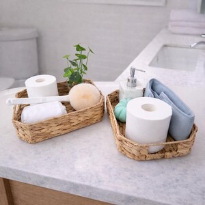 May include: Two woven wicker baskets on a bathroom countertop. One basket holds a roll of toilet paper, a white washcloth, a brush, and a beige sponge. The other basket contains a roll of toilet paper, a blue washcloth, a soap dispenser, and a green decorative item.