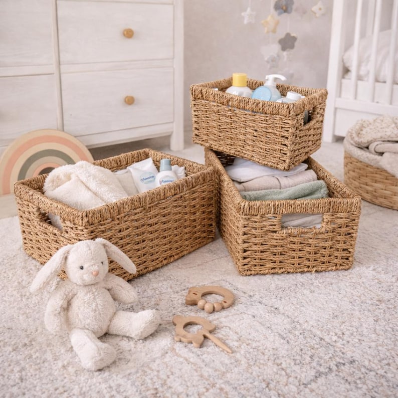 Peut inclure: Trois paniers de rangement en osier tress&eacute;, empil&eacute;s et remplis d'articles pour b&eacute;b&eacute;s, sont pos&eacute;s sur un tapis de couleur claire. Un lapin en peluche et des jouets en bois sont devant les paniers. Les paniers contiennent des produits et des serviettes pour b&eacute;b&eacute;s.