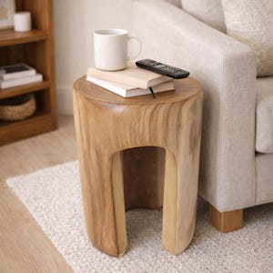 May include: A natural wood side table with a unique, rounded design. The table has a hollowed-out center and is topped with a white mug, two books, and a black remote control. The table is next to a beige sofa.