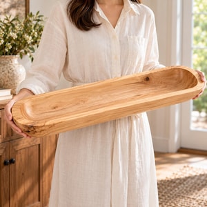 May include: A long, narrow, oval-shaped wooden dough bowl with a smooth, light brown finish and visible natural wood grain patterns. The bowl features a deep, hollowed-out center and a thick, sturdy rim with a rustic, textured exterior surface.