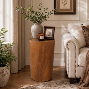 May include: A cylindrical wooden side table with a smooth, light brown grain finish. The table supports a textured, light gray ceramic vase filled with green leafy branches, a small dark-framed picture, and a glass candle with a lit wick.