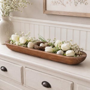 May include: A long, oval-shaped wooden dough bowl filled with white tulip flowers, small green sprigs, and several speckled eggs nestled within a small woven twig nest. The bowl has a smooth, dark brown finish and sits horizontally.