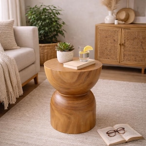 May include: A light brown wooden side table with a unique hourglass shape. The table has a round top with a succulent plant in a white pot, a book, and a glass of water with a lemon slice. The table is on a light-colored rug.