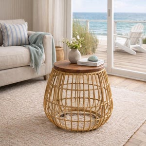 May include: A round accent table with a woven, light brown base and a dark wood top. The table is in a living room setting, with a sofa, rug, and ocean view in the background. A vase of flowers and a book are on the table.