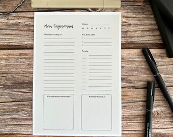 May include: A white daily planner with German text, including "Mein Tagesramn" and sections for notes and appointments. Two black pens, a pink and a beige highlighter, and a black notebook are also visible on a wooden surface.