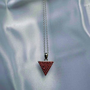May include: A silver necklace featuring a triangular pendant. The pendant is adorned with numerous small, red gemstones. The chain is fine and suspends the pendant. The backdrop is a white, silky material.