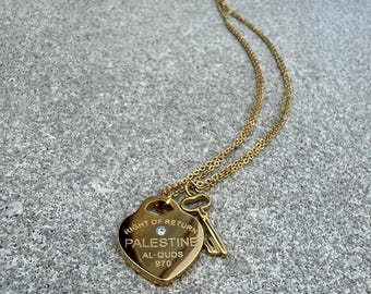 May include: A gold-colored necklace with a heart-shaped pendant and a key charm. The pendant features the text "RIGHT OF RETURN PALESTINE AL-QUDS 970" and a small gemstone. The necklace is on a gray surface.