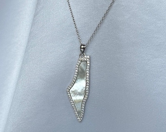 May include: A silver necklace with a pendant shaped like a map outline. The pendant is filled with a pearlescent material and is outlined with small, clear gemstones. The necklace hangs against a light gray background.