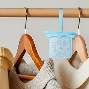 May include: A blue fabric hanging air freshener with a floral design and the text "Lavender & Cedarwood" hanging on a wooden clothes rack with two jumpers on hangers.