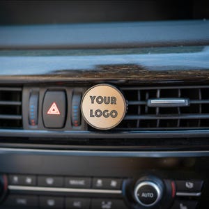 May include: A round, gold-colored car air freshener with the text "YOUR LOGO" printed on it. The air freshener is attached to a car's air vent.