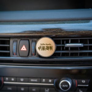 May include: A round, tan car air vent freshener with Japanese text. The freshener is mounted on a car's dashboard, near the hazard lights. The dashboard is black with wood grain accents.