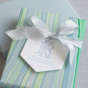 May include: A blue and green striped gift box with a white tag that says "Katherine Ricketson" and a blue monogram. The box is tied with a blue ribbon.