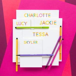 May include: Five white notepads with colorful names written on them: Charlotte, Lucy, Jackie, Tessa, and Skyler. The notepads are stacked on top of each other and surrounded by colorful pens.