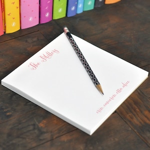 May include: A white notepad with the text "The Millers" printed in pink cursive. A black pencil with a pink and white patterned design is on top of the notepad. The notepad has a gold edge and the text "even. calmer. etc. alive" printed in pink cursive on the bottom edge.