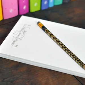 May include: A white notepad with the text "Ashley and Darryl" in a cursive font. A black and gold pencil is lying on top of the notepad.
