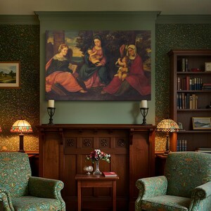 May include: A traditional living room with a large painting above a wooden fireplace. Two green patterned armchairs are positioned either side of the fireplace, with matching lamps. A small side table holds a vase of flowers.