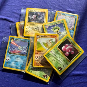 May include: A collection of Pokemon trading cards featuring various characters, including Dark Golduck, Lt. Surge's Magnemite, Erika's Exeggutor, Seadra, and Marowak. The cards are in protective plastic sleeves and are arranged in a pile.