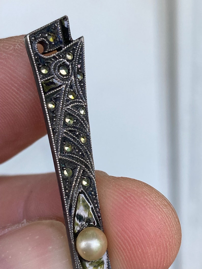 May include: A silver brooch with a geometric design and a pearl accent. The brooch is decorated with small, sparkling stones.