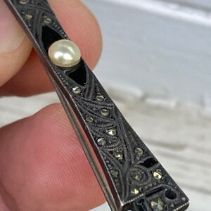 May include: A silver brooch with a black enamel design and a pearl in the center. The brooch is decorated with marcasite stones.