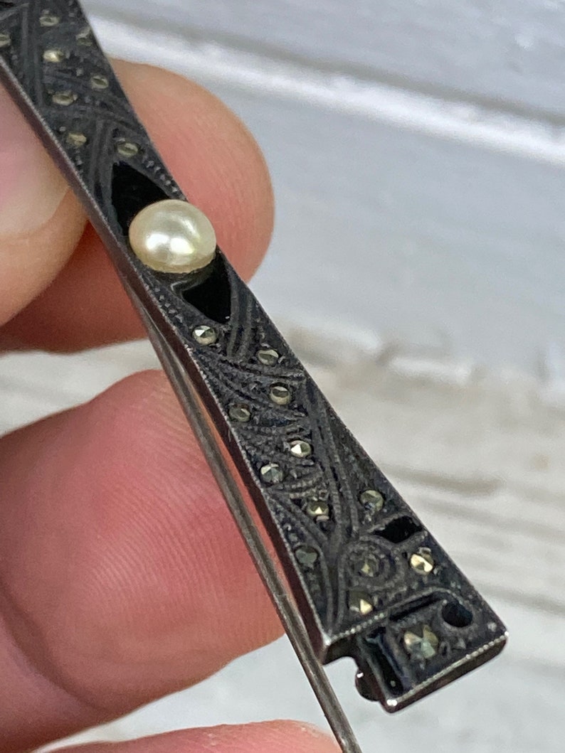 May include: A silver brooch with a black enamel design and a pearl in the center. The brooch is decorated with marcasite stones.