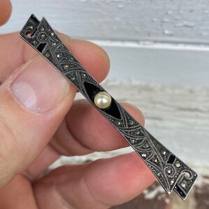 May include: A silver and black bar brooch with a pearl in the center. The brooch is decorated with a geometric design and black enamel.