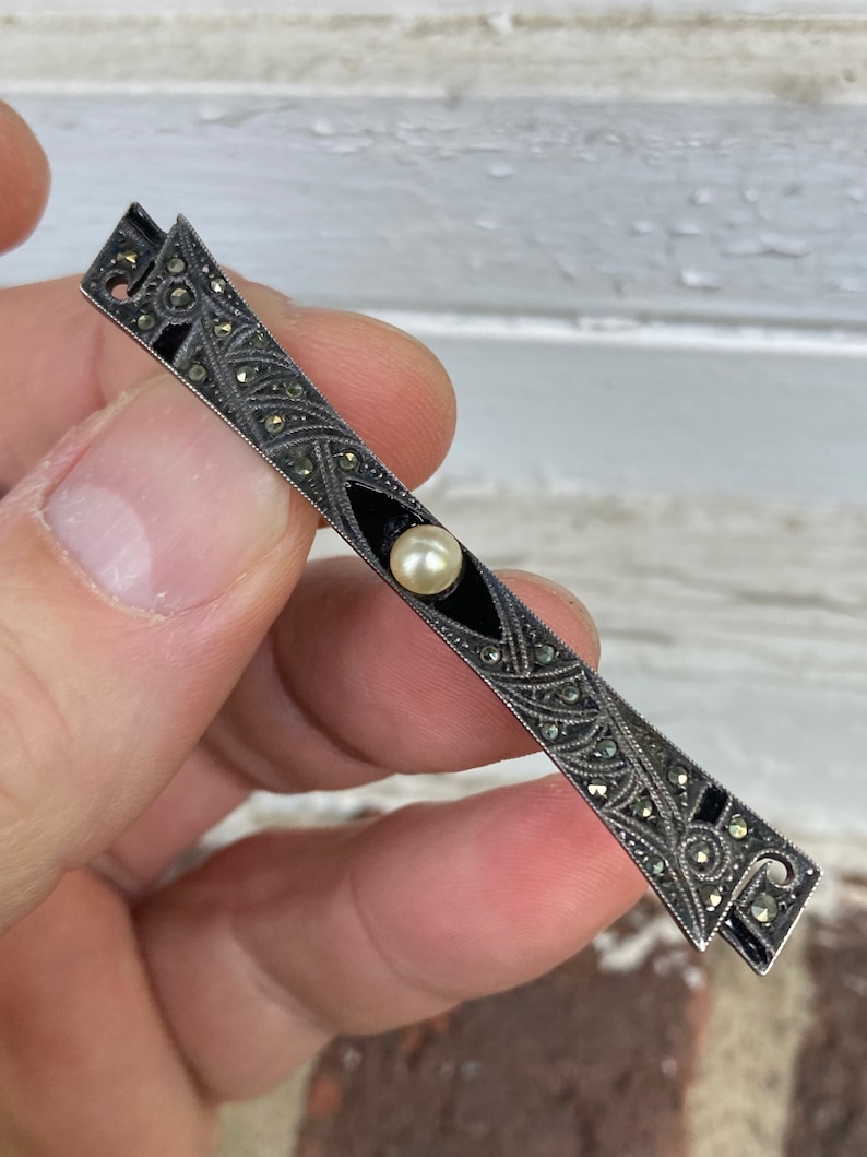 May include: A silver and black bar brooch with a pearl in the center. The brooch is decorated with a geometric design and black enamel.