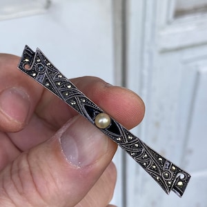May include: A silver brooch with a black enamel design and a pearl in the center. The brooch is decorated with marcasite stones.