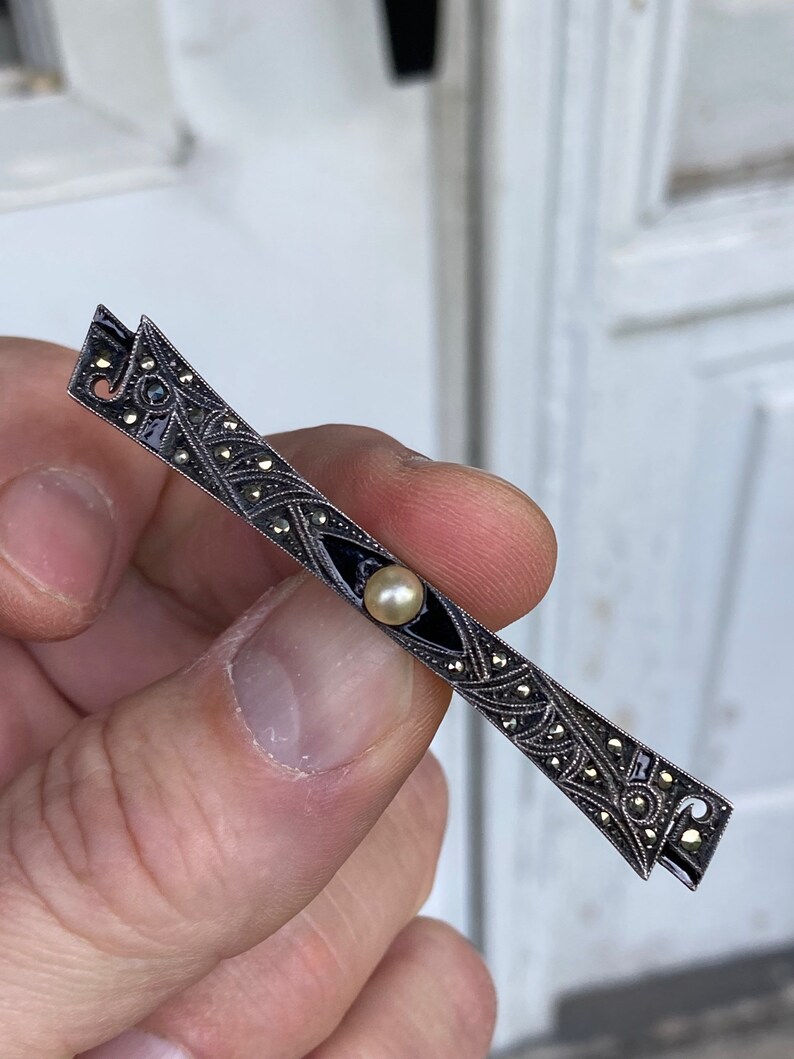 May include: A silver brooch with a black enamel design and a pearl in the center. The brooch is decorated with marcasite stones.