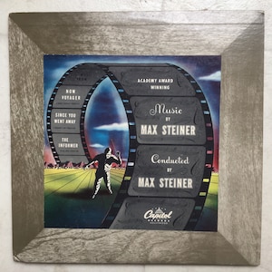 May include: A vintage record album cover featuring a film reel with the text "Music by Max Steiner" and "Conducted by Max Steiner". The album cover is from Capitol Records, Hollywood. The album cover also features a man in a white suit conducting an orchestra.