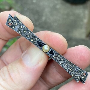 May include: A silver bar pin with a black enamel center and a single pearl. The pin is decorated with marcasite stones.